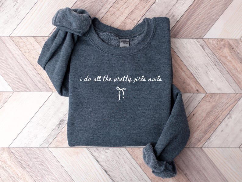 May include: A dark gray sweatshirt with the phrase "i do all the pretty girls nails" in white script, along with a small bow graphic. The sweatshirt is folded on a light brown and white wooden surface.