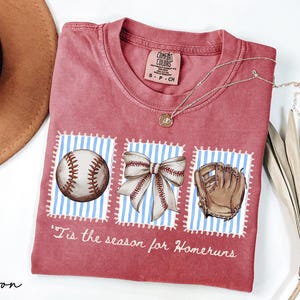 May include: Crimson-colored t-shirt with baseball-themed graphics. The shirt features a baseball, a bow, and a baseball glove on blue and white striped backgrounds. The text "'Tis the season for Homeruns" is printed on the shirt.