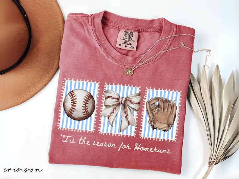 May include: Crimson-colored t-shirt with baseball-themed graphics. The shirt features a baseball, a bow, and a baseball glove on blue and white striped backgrounds. The text "'Tis the season for Homeruns" is printed on the shirt.