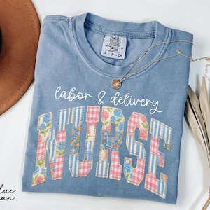 May include: Blue jean Comfort Colors t-shirt with the words "labor & delivery nurse" in white script and patchwork letters. The letters are decorated with floral and gingham patterns. A gold necklace is draped across the shirt.