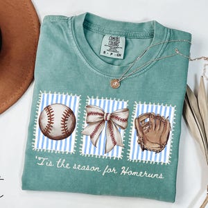 May include: Light green t-shirt with baseball-themed graphics. The shirt features a baseball, a bow, and a baseball glove on blue and white striped backgrounds. The text "'Tis the season for Homeruns" is printed below the graphics.