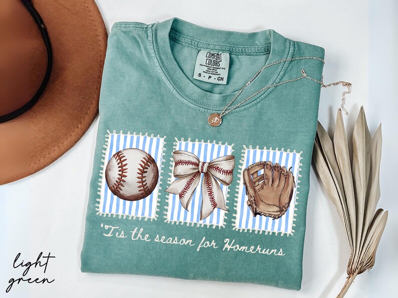 May include: Light green t-shirt with baseball-themed graphics. The shirt features a baseball, a bow, and a baseball glove on blue and white striped backgrounds. The text "'Tis the season for Homeruns" is printed below the graphics.