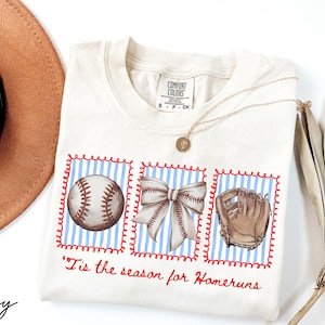 May include: Ivory-colored t-shirt with a baseball theme. The shirt features three framed illustrations: a baseball, a bow, and a baseball glove, set against a blue and white striped background. The phrase "'Tis the season for Homeruns" is written in red.