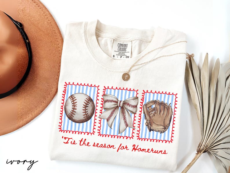 May include: Ivory-colored t-shirt with a baseball theme. The shirt features three framed illustrations: a baseball, a bow, and a baseball glove, set against a blue and white striped background. The phrase "'Tis the season for Homeruns" is written in red.