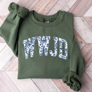 May include: An olive green sweatshirt with the letters "WWJD" in a white and blue floral pattern across the chest. The sweatshirt is folded on a light wood-grain surface.