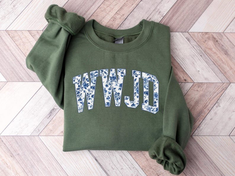 May include: An olive green sweatshirt with the letters "WWJD" in a white and blue floral pattern across the chest. The sweatshirt is folded on a light wood-grain surface.