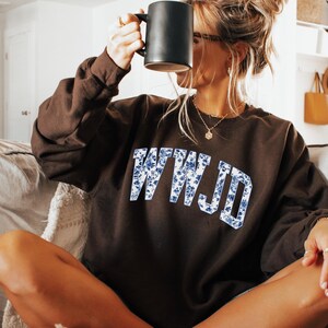 May include: A dark brown sweatshirt with the letters "WWJD" in a blue floral pattern. The person in the image is holding a black mug. The sweatshirt is a casual style.
