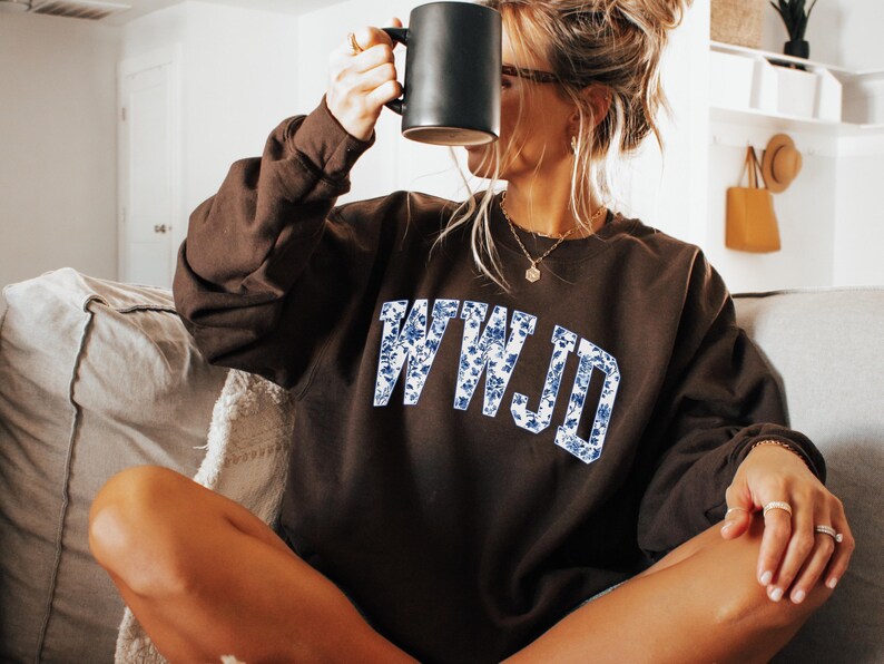 May include: A dark brown sweatshirt with the letters "WWJD" in a blue floral pattern. The person in the image is holding a black mug. The sweatshirt is a casual style.