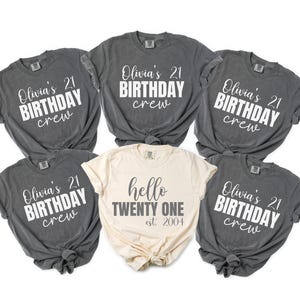 Op de afbeelding: Zes t-shirts. Vijf zijn donkergrijs met de witte tekst "Olivia's 21 BIRTHDAY crew". De zesde, cr&egrave;mekleurige shirt heeft de tekst "hello TWENTY ONE est. 2004".
