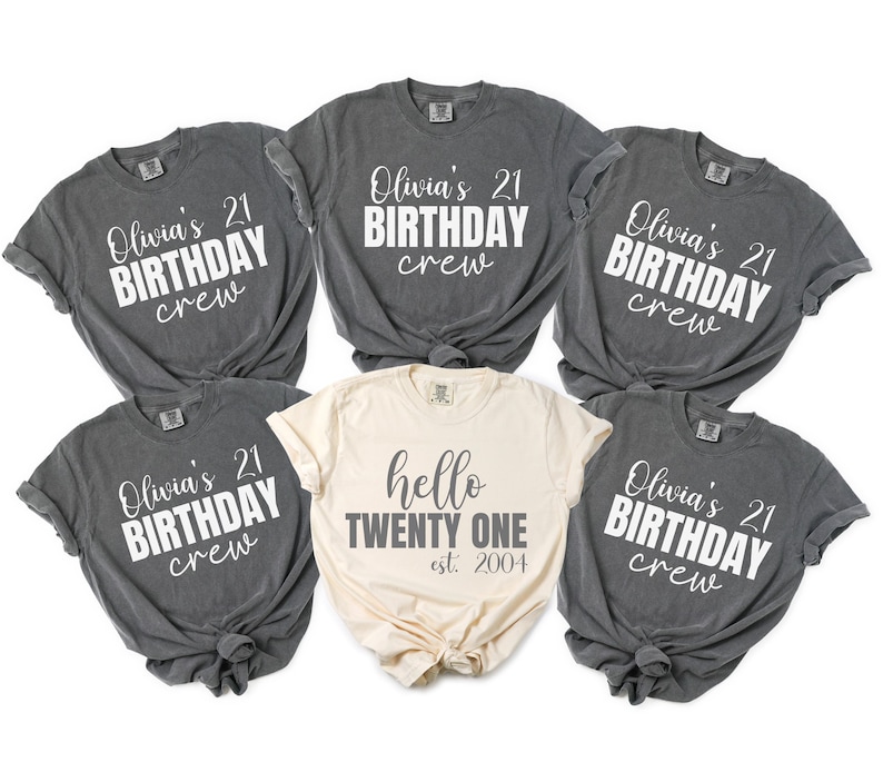 Op de afbeelding: Zes t-shirts. Vijf zijn donkergrijs met de witte tekst "Olivia's 21 BIRTHDAY crew". De zesde, cr&egrave;mekleurige shirt heeft de tekst "hello TWENTY ONE est. 2004".