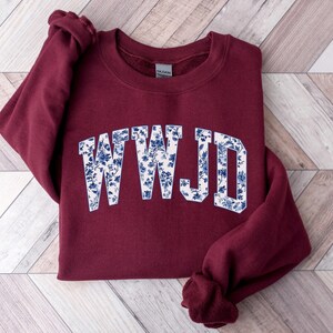 May include: A maroon sweatshirt with the letters "WWJD" in a white font filled with a blue floral pattern. The sweatshirt is folded on a light wood-grain surface.