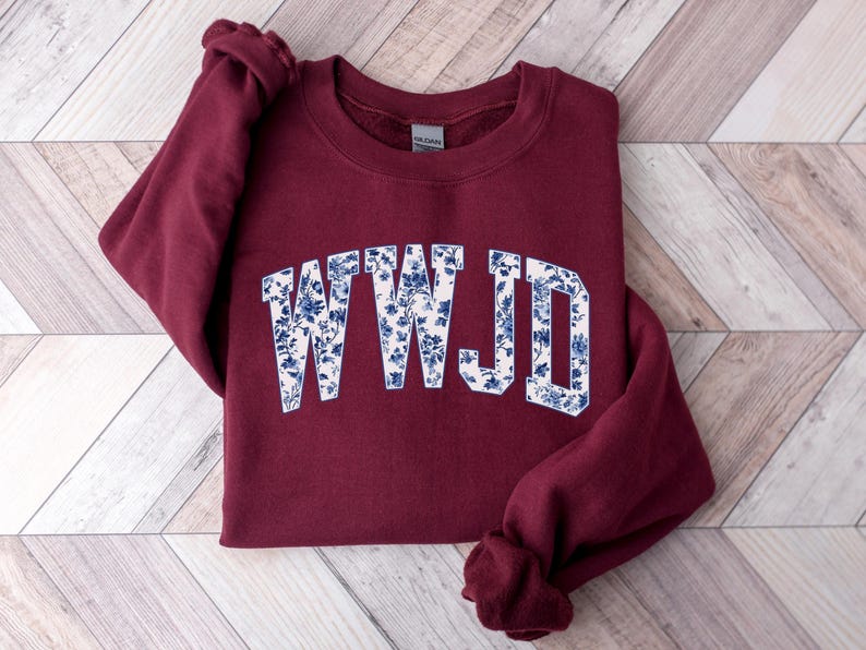 May include: A maroon sweatshirt with the letters "WWJD" in a white font filled with a blue floral pattern. The sweatshirt is folded on a light wood-grain surface.