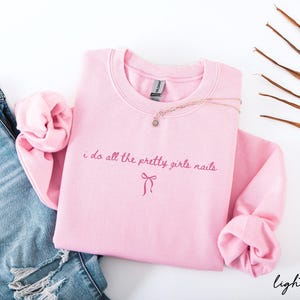 May include: A light pink sweatshirt with the text "i do all the pretty girls nails" in a cursive font. A small bow graphic is below the text. A gold necklace and a pair of blue jeans are next to the sweatshirt.