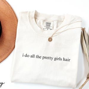 Może przedstawiać: Kremowa koszulka z napisem "i do all the pretty girls hair" w kolorze czarnym. Złoty naszyjnik z wisiorkiem spoczywa na koszulce. Brązowy kapelusz i suszona roślina dekoracyjna r&oacute;wnież znajdują się na zdjęciu.