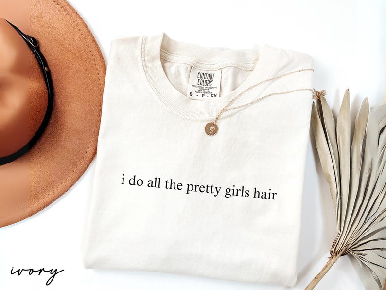 Może przedstawiać: Kremowa koszulka z napisem "i do all the pretty girls hair" w kolorze czarnym. Złoty naszyjnik z wisiorkiem spoczywa na koszulce. Brązowy kapelusz i suszona roślina dekoracyjna r&oacute;wnież znajdują się na zdjęciu.