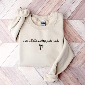 May include: A cream-colored sweatshirt with the text "i do all the pretty girls nails" in black script, with a bow graphic underneath. The sweatshirt has a crew neck and long sleeves.