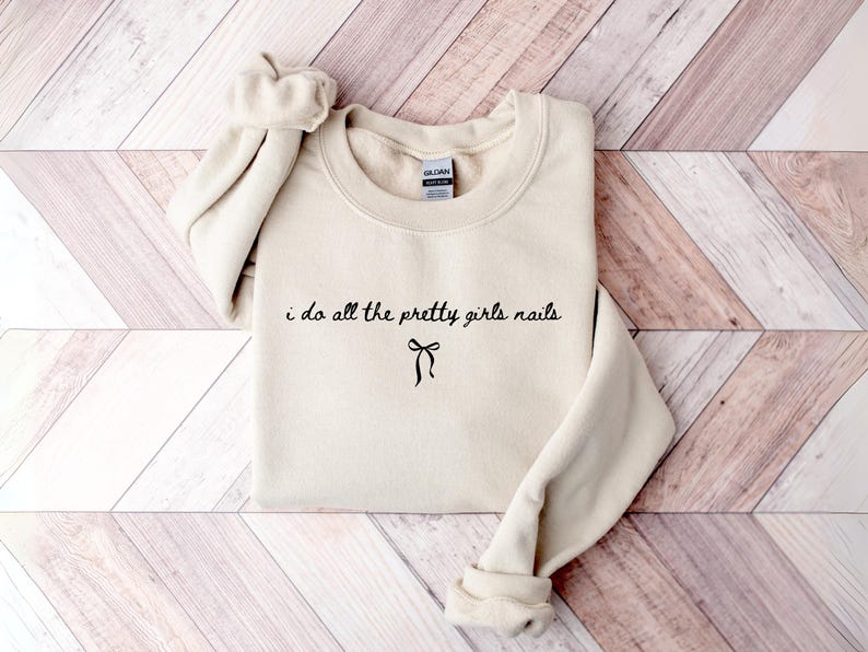 May include: A cream-colored sweatshirt with the text "i do all the pretty girls nails" in black script, with a bow graphic underneath. The sweatshirt has a crew neck and long sleeves.