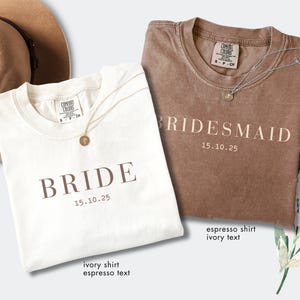 May include: Two t-shirts, one ivory and one espresso, with the words "BRIDE" and "BRIDESMAID" printed on them. Each shirt has a gold necklace. The date "15.10.25" is printed below the words. A tan hat is in the background.