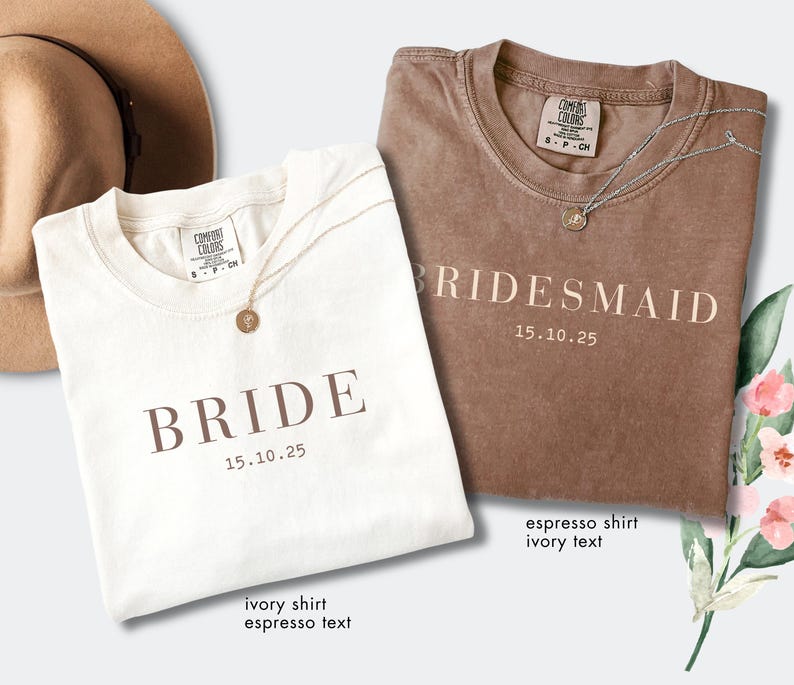 May include: Two t-shirts, one ivory and one espresso, with the words "BRIDE" and "BRIDESMAID" printed on them. Each shirt has a gold necklace. The date "15.10.25" is printed below the words. A tan hat is in the background.