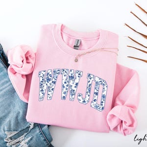 May include: A light pink sweatshirt with the text "WWJD" in a blue floral design. The sweatshirt is folded with the sleeves tucked in. A gold necklace with a small pendant is on top of the sweatshirt. Blue jeans are on the left.
