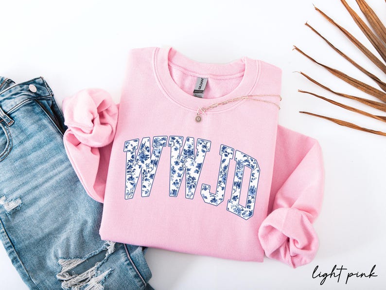 May include: A light pink sweatshirt with the text "WWJD" in a blue floral design. The sweatshirt is folded with the sleeves tucked in. A gold necklace with a small pendant is on top of the sweatshirt. Blue jeans are on the left.