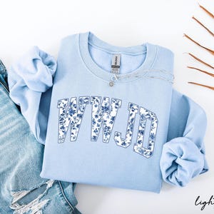 May include: Light blue sweatshirt with the text "WWJD" in a floral design. The sweatshirt is paired with light wash denim jeans and a silver necklace. The words "light blue" are written in script at the bottom right.
