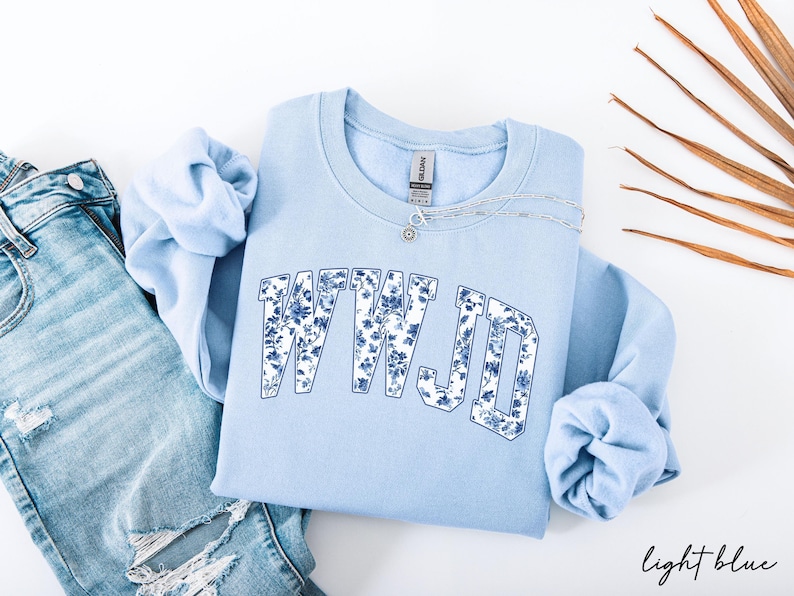 May include: Light blue sweatshirt with the text "WWJD" in a floral design. The sweatshirt is paired with light wash denim jeans and a silver necklace. The words "light blue" are written in script at the bottom right.