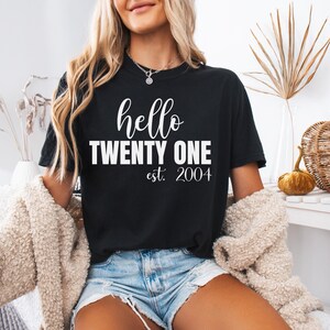 Op de afbeelding: Zwart T-shirt met de tekst "hello TWENTY ONE est. 2004" in witte letters en blokletters. Het shirt heeft een ronde hals. Het model draagt een blauwe spijkerbroek en een cr&egrave;mekleurig vest.