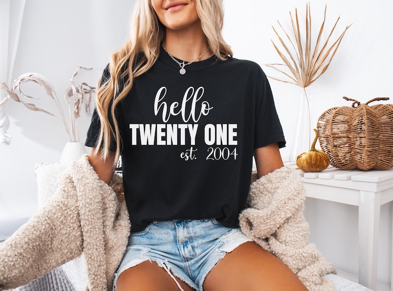 Op de afbeelding: Zwart T-shirt met de tekst "hello TWENTY ONE est. 2004" in witte letters en blokletters. Het shirt heeft een ronde hals. Het model draagt een blauwe spijkerbroek en een cr&egrave;mekleurig vest.