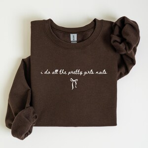 May include: A dark brown sweatshirt with the text "i do all the pretty girls nails" in white script. A white bow graphic is below the text. The sweatshirt is folded, with the sleeves visible.