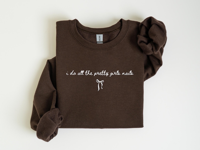 May include: A dark brown sweatshirt with the text "i do all the pretty girls nails" in white script. A white bow graphic is below the text. The sweatshirt is folded, with the sleeves visible.
