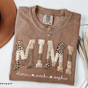 May include: Espresso-colored t-shirt with the word "MIMI" appliqued in a patchwork design featuring leopard print and patterned fabrics. The names "olivia", "noah", and "sophie" are printed below. A silver necklace and a brown hat are also visible.