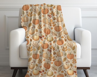 May include: A cozy throw blanket draped over a white armchair. The blanket features a repeating pattern of orange pumpkins, fall leaves, and berries in shades of orange, brown, and cream, creating a warm, autumnal theme.