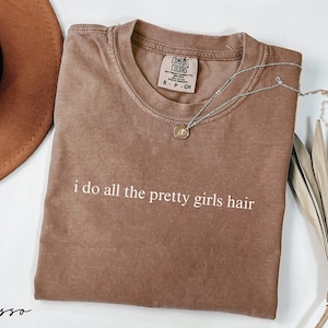 Espresso brown Comfort Colors® shirt with minimalist white text design that reads "i do all the pretty girls hair", styled with necklace and hat