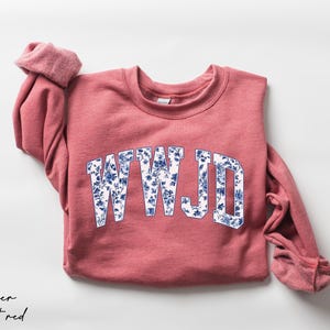May include: A heather scarlet red sweatshirt with the letters "WWJD" in a white and blue floral design. The sleeves are rolled up, and the sweatshirt is on a white background.