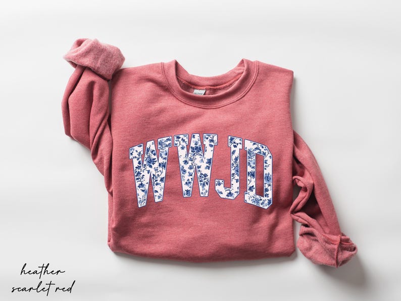May include: A heather scarlet red sweatshirt with the letters "WWJD" in a white and blue floral design. The sleeves are rolled up, and the sweatshirt is on a white background.
