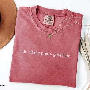 Może przedstawiać: Karmazynowa koszulka z białym napisem "i do all the pretty girls hair". Widoczny jest r&oacute;wnież złoty naszyjnik z wisiorkiem i brązowy kapelusz. Po prawej stronie znajduje się dekoracyjna suszona roślina.
