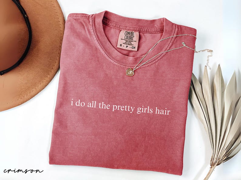 Może przedstawiać: Karmazynowa koszulka z białym napisem "i do all the pretty girls hair". Widoczny jest r&oacute;wnież złoty naszyjnik z wisiorkiem i brązowy kapelusz. Po prawej stronie znajduje się dekoracyjna suszona roślina.