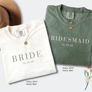May include: Two t-shirts, one ivory and one moss green, are displayed. The ivory shirt reads "BRIDE" with the date "15.10.25" in moss text. The green shirt says "BRIDESMAID" with the same date in ivory text. Both shirts have a necklace.