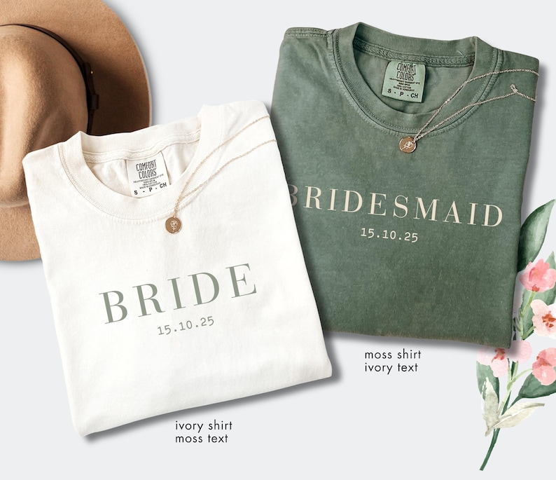 May include: Two t-shirts, one ivory and one moss green, are displayed. The ivory shirt reads "BRIDE" with the date "15.10.25" in moss text. The green shirt says "BRIDESMAID" with the same date in ivory text. Both shirts have a necklace.