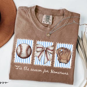 May include: Espresso-colored t-shirt with a baseball theme. The shirt features three postage stamp-like designs with a baseball, bow, and glove. The text "'Tis the season for Homeruns" is printed below the designs. A silver necklace and a hat are also visible.