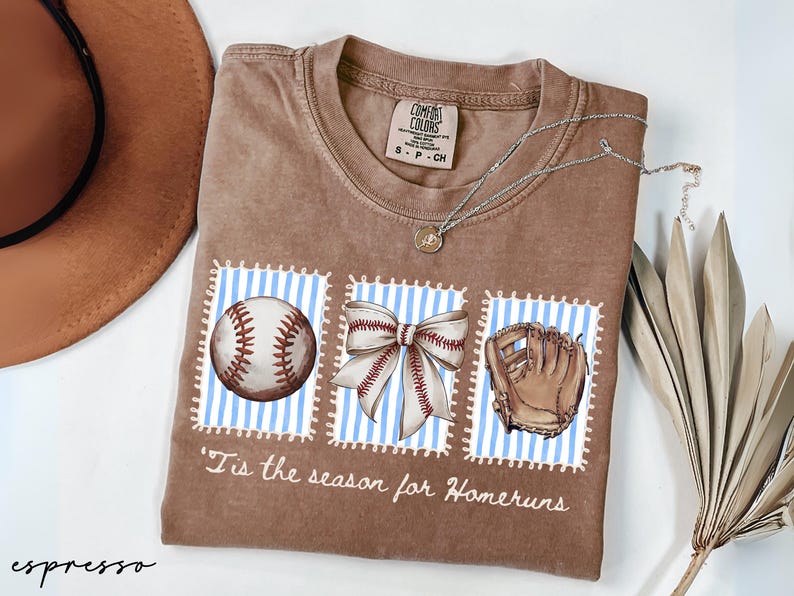 May include: Espresso-colored t-shirt with a baseball theme. The shirt features three postage stamp-like designs with a baseball, bow, and glove. The text "'Tis the season for Homeruns" is printed below the designs. A silver necklace and a hat are also visible.