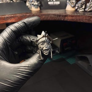 May include: A silver-colored ring featuring a detailed, smiling face with a helmet. The ring has intricate detailing, including the face's features and the helmet's design. The ring is held by a gloved hand, showcasing the craftsmanship.