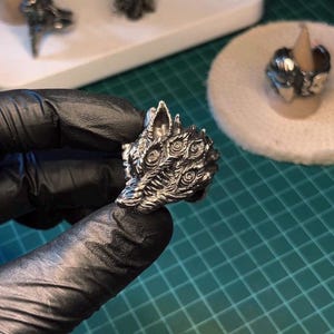 Puede incluir: Un anillo plateado con un diseño detallado de cabeza de lobo. El anillo muestra intrincados detalles del pelaje, los ojos y el hocico del lobo. Otros anillos son visibles en el fondo, lo que sugiere una colección o exhibición de joyas similares.