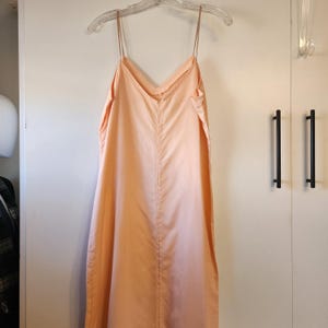 May include: A peach-colored slip dress with thin spaghetti straps hangs on a clear hanger. The dress has a V-neckline and a loose, flowing silhouette. The fabric appears to be a lightweight material, suitable for warm weather.