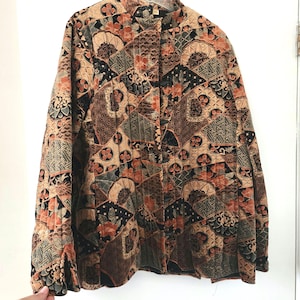 May include: A quilted jacket with a mandarin collar and a patchwork design. The jacket features a mix of geometric and floral patterns in shades of brown, black, orange, and beige. The jacket has long sleeves and a button-up front.