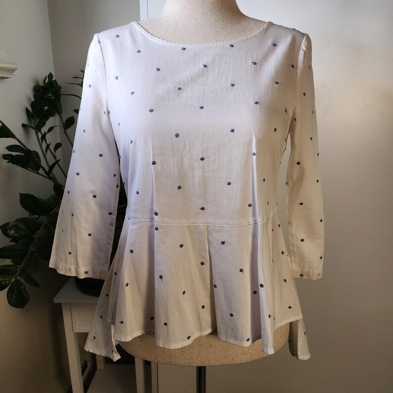 May include: White blouse with a blue polka dot pattern. The blouse has a ruffled hem and a slightly asymmetrical cut.