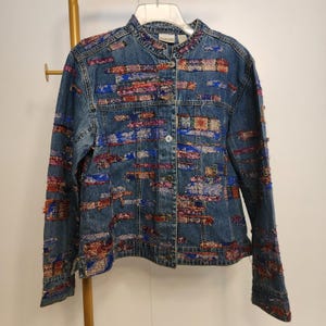May include: Blue denim jacket with colorful embroidered patches. The jacket features a mandarin collar, button-up front, and long sleeves. The patches are in shades of red, blue, and purple.