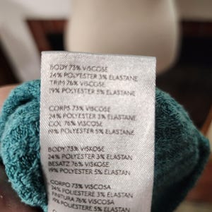 Puede incluir: Primer plano de una tela turquesa con una etiqueta blanca. La etiqueta indica la composici&oacute;n del tejido: 73% viscosa, 24% poli&eacute;ster, 3% elastano, y 76% viscosa, 19% poli&eacute;ster, 5% elastano. El texto se repite.