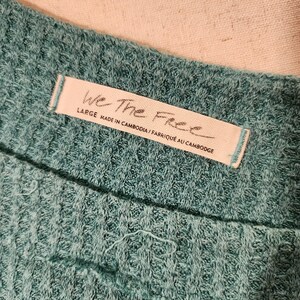 Puede incluir: Primer plano de una tela de punto de gofre de color verde azulado. Una etiqueta blanca con el texto "We The Free" est&aacute; cosida a la tela. La etiqueta tambi&eacute;n incluye la talla "LARGE" y el texto "MADE IN CAMBODIA / FABRIQU&Eacute; AU CAMBODGE".